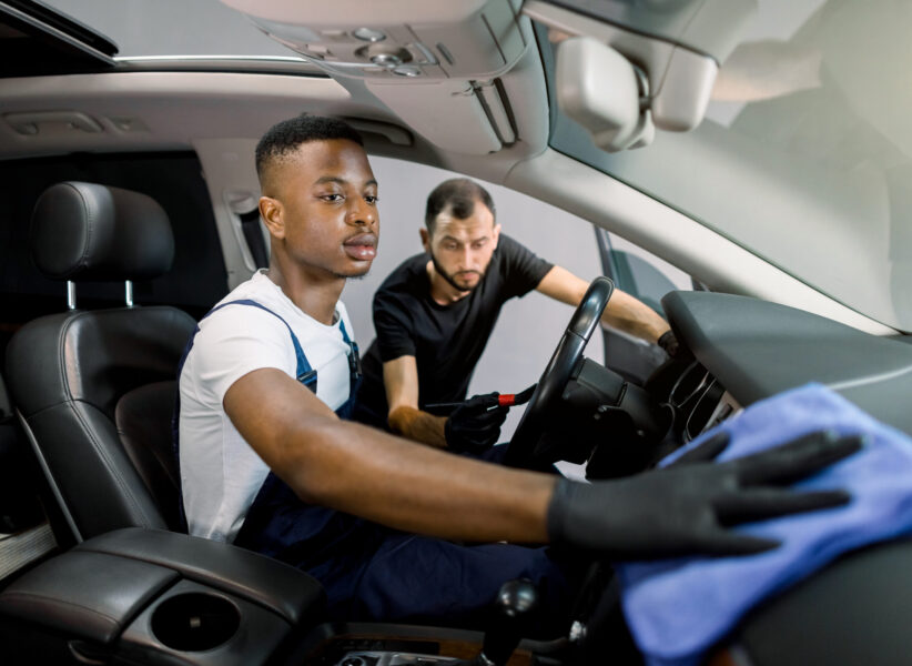 professional car cleaning and detailing concept. Two multiracial male workers in uniforms, washing car interior with brush, microfiber and detergent at service station. Focus on hand of African man.