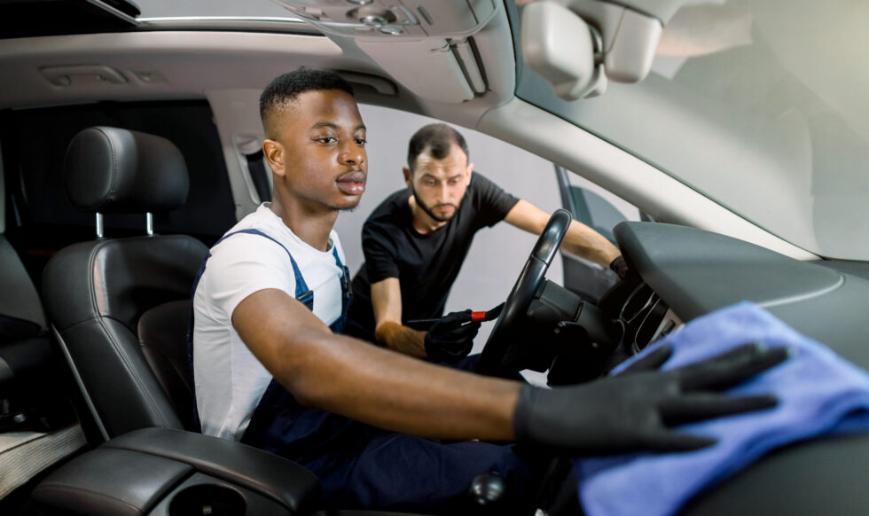 professional car cleaning and detailing concept. Two multiracial male workers in uniforms, washing car interior with brush, microfiber and detergent at service station. Focus on hand of African man.