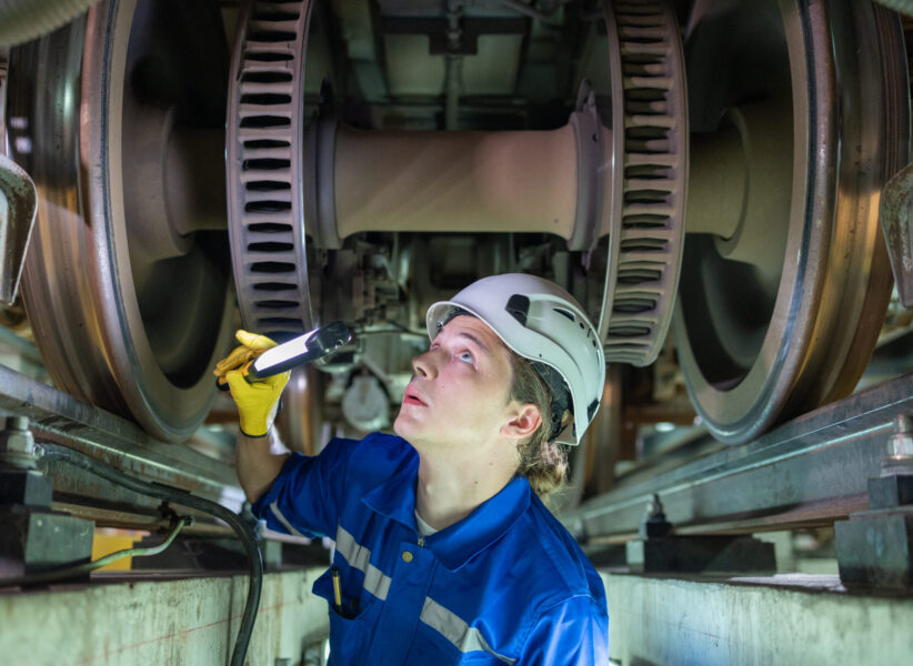 Focused maintenance worker in a helmet and blue uniform using a flashlight to inspect the wheel assembly of a train.