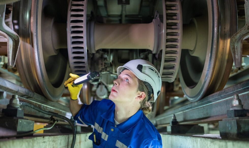 Maintenance Worker Examining Train Wheels with Torch Focused maintenance worker in a helmet and blue uniform using a flashlight to inspect the wheel assembly of a train.